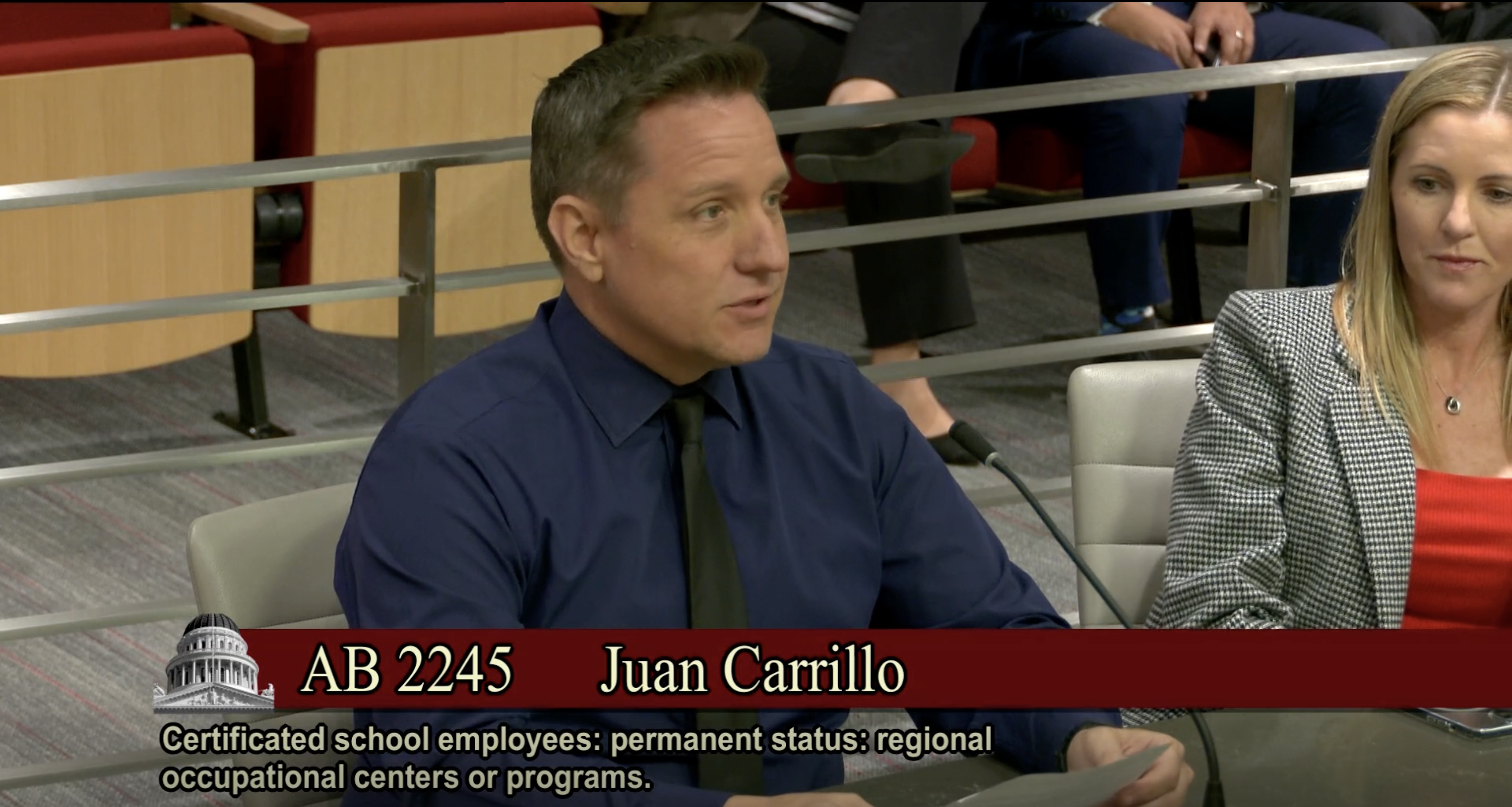 A teacher at a microphone speaks to the state board of education. Label shows that this is a hearing for AB 2245, sponsored by Juan Carrillo.