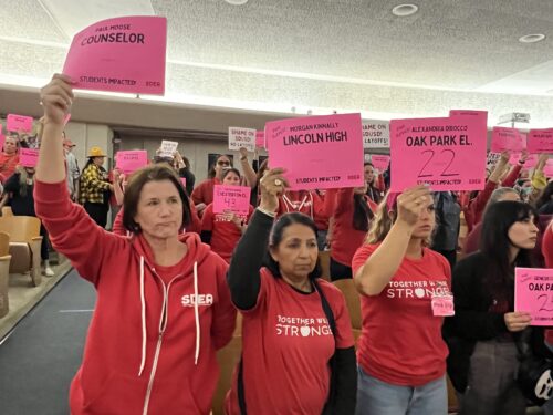 Educators wearing red union shirts and serious expressions hold up pink slips